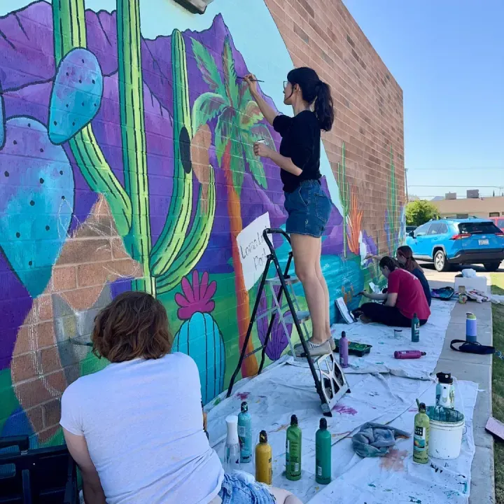 Brigitte De Marco on a ladder painting a desert mural with community helpers
